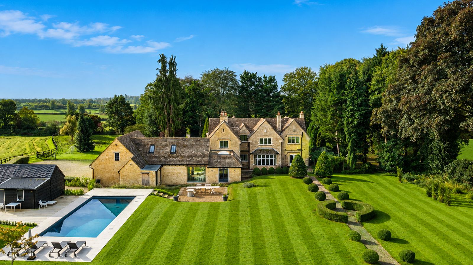 Tormead House from the air: the Cotswold stone house and barn, the heated pool on its stone terrace, striped lawns and clipped topiary, with open Cotswold countryside beyond.