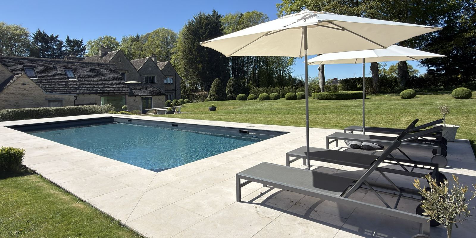 The heated outdoor pool at Tormead House, edged in pale stone with loungers and parasols, the Cotswold stone house and clipped topiary beyond.