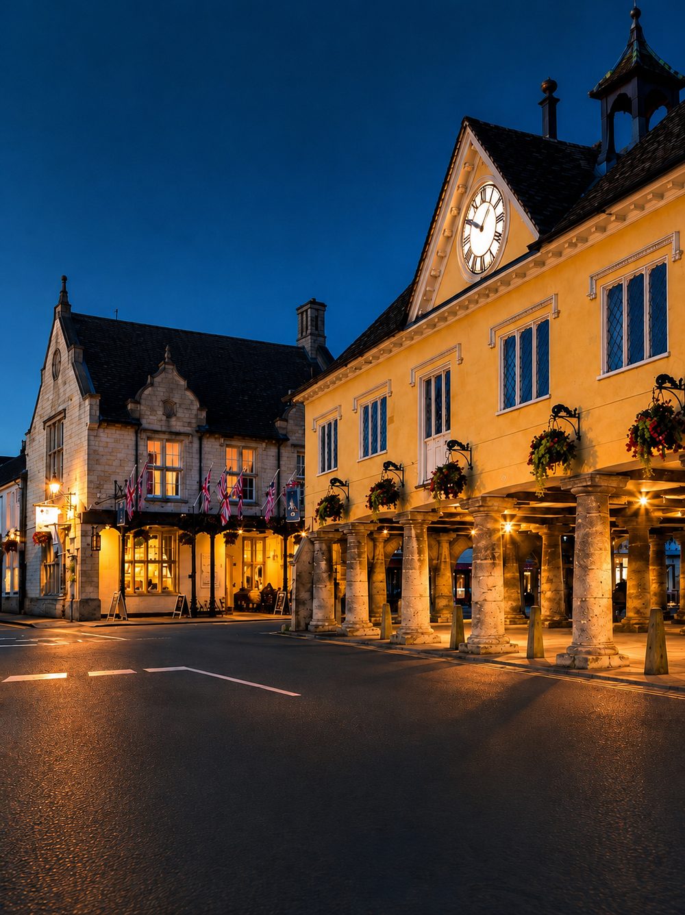 The Market House at Tetbury at dusk, its clock tower lit and the ground floor open between three rows of stone pillars, with the lit windows of Long Street beyond.