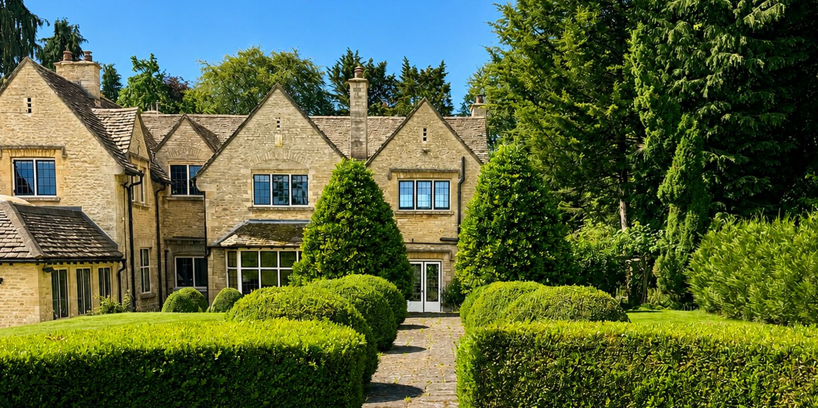 Tormead House seen from the garden: a Cotswold stone house with three gables and stone mullioned windows, behind clipped box hedging and a flagstone path.